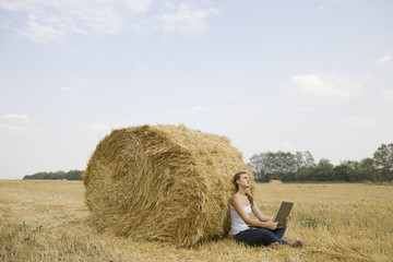 Naklejka na ściany i meble Side view of a young woman with laptop sitting against hay bale in field