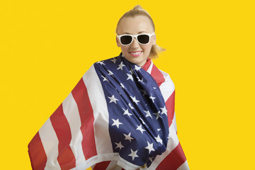 Portrait of happy young woman wrapped in American flag over yellow background