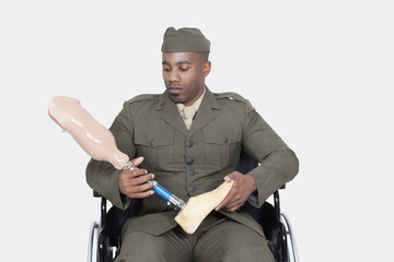 Sad US military officer in wheelchair holding prosthesis foot over gray background