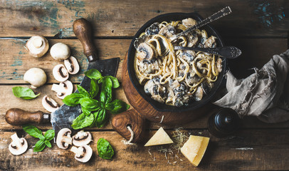 Italian style dinner. Creamy mushroom pasta spaghetti in cast iron pan on wooden boards with Parmesan cheese, fresh basil leaves and pepper over old rustic background. Top view, horizontal composition