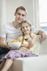 Obraz na płótnie Canvas Portrait of father and daughter watching TV with bowl full of wheel shape snack pellets