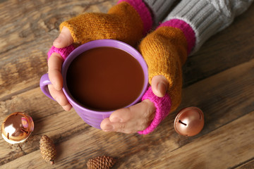 Fototapeta na wymiar Female hands in mittens holding cup of coffee on wooden background