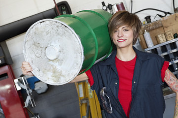 Portrait of a happy young female mechanic carrying oil drum on shoulder in car workshop