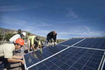 Group of multiethnic engineers lifting heavy solar panel against blue sky