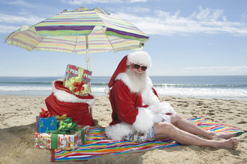 Portrait of a happy man dressed in Santa Claus outfit with gifts sitting under parasol on beach