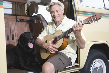 Portrait of a happy senior man sits in his campervan with a guitar and his pet dog