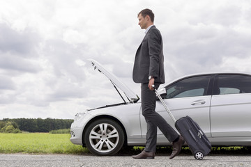 Fototapeta na wymiar Full length side view of young businessman with luggage walking by broken down car at countryside