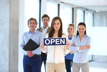 Portrait of young businesswoman holding open sign with team behind at office