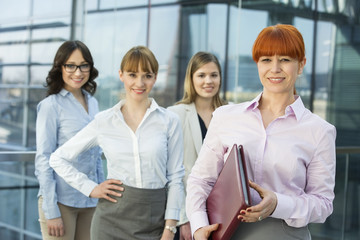 Portrait of confident businesswoman holding folder with female colleagues standing in background