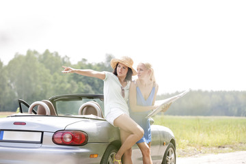 Naklejka premium Woman showing something to female friend with map while leaning on convertible