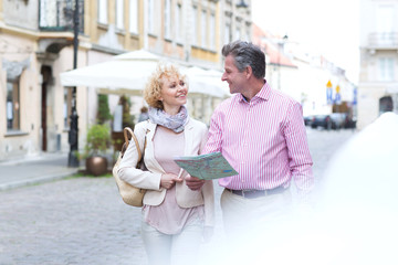 Smiling middle-aged couple with map looking at each other while walking in city