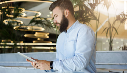 Side view. Young bearded businessman wearing blue shirt uses tablet computer. Man checks email on digital tablet, browsing internet, chatting. Lights in shape of circles in soft focus in background.