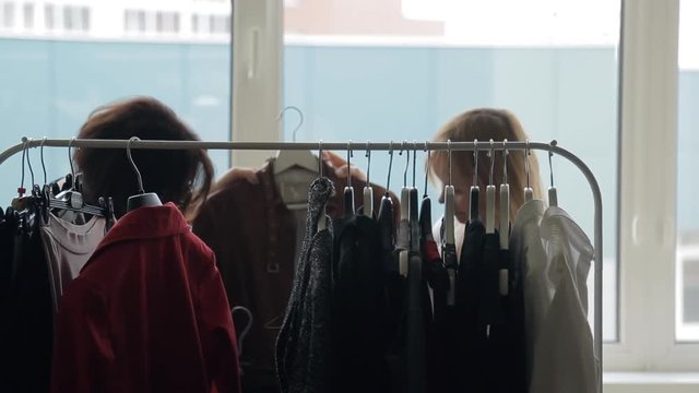 Two women discuss fashion trends, view clothes on rack with hangers.