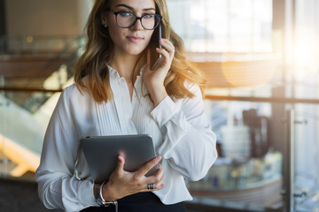 Fototapeta premium Cropped image.Front view. Young business woman in glasses and white shirt, standing in room with modern interior and talking on cellphone while holding tablet computer in her hand. Girl using gadget.