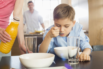 Midsection of woman with juice bottle standing by son having breakfast with man in background