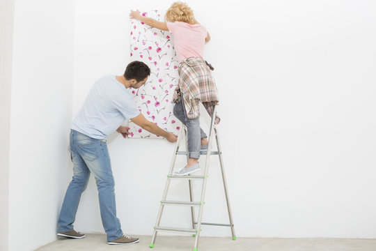 Full-length rear view of couple applying wallpaper to wall