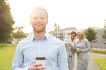 Portrait of happy businessman holding disposable cup with colleagues standing in background