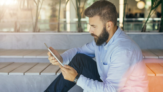 Side view. Young bearded businessman in blue shirt is sitting in room on bench and using tablet computer. Man holding digital tablet and looking at screen.Man using gadget, checking email.Film effect.