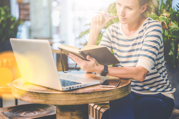 Young woman in striped t-shirt sitting at round wooden table in cafe and holding notebook and pen. In front of her is laptop, smartphone. Freelancer working outside home. Student learning online.