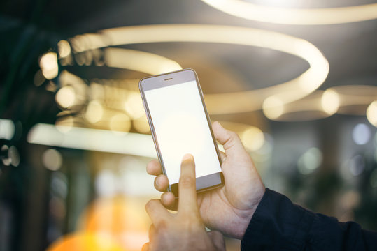 Close-up of a smartphone with a blank screen in male hands. Male finger on the screen of the gadget. Abstract lights in soft focus in the background. Mock up. Advertising space.