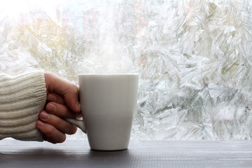 Obraz na płótnie Canvas warming in winter/ white mug with a hot drink in hand dressed in a sweater against the window with frosty patterns