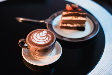 Cappuchino or latte coffe in a white cup on with a cake on a black table.