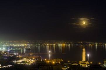 Night view of the city of Baku - the capital of the Republic of Azerbaijan with full moon in soft clouds over dark water with reflections on it