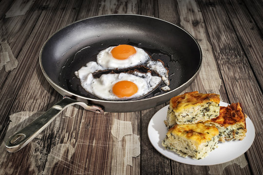 Sunny Side Up Fried Eggs in Old Teflon Frying Pan and Plateful of Cheese Pie Gibanica Slices Set on Old Cracked Flaky Wooden Table