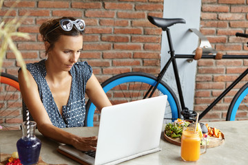 Fototapeta na wymiar Casual businesswoman wearing shades on her head keyboarding on laptop, checking email during lunch on her day-off, sitting in front of open generic notebook with serious and concentrated expression