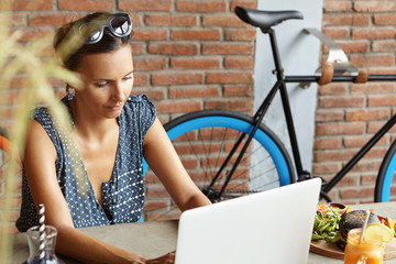 Fototapeta na wymiar Beautiful female freelancer using laptop computer for remote work, sitting at table with food, looking at screen with faint smile, enjoying free wi-fi, red brick wall and bicycle on background