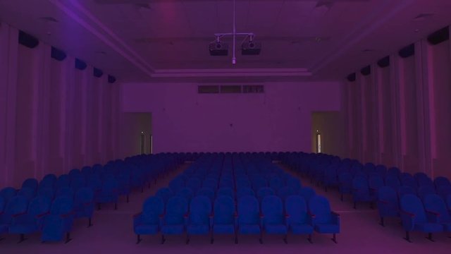 Modern concert hall with a lot of seats. Room off the main light and only spotlight with colored lights illuminate it. amera moves away from the seat in the hall