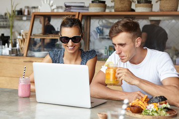 People, technology and leisure concept. Cute couple having fun, sitting at cafe in front of laptop computer: man sipping orange juice and woman in sunglasses absorbed in watching something on notebook