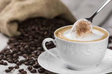 Cappuccino Coffee Cup and Spoon / Composition of Cup of Cappuccino and spoon with foam, sack of coffee beans on white wooden table