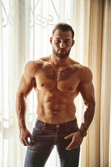Fototapeta premium Portrait of sexy shirtless muscular man next to window curtains during the day, wearing only jeans