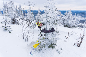 Young beautiful woman in bright yellow cap, ski mask, on snowboard smiles and embraces snow-covered fir-tree at snow mountain top