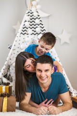 Mother, father and son lying on the floor with Christmas tree and Christmas presents, play, joke, laugh, spend time together.