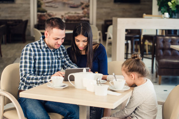 Family with child in cafe. Mother and father watching on tablet pc while girl waiting