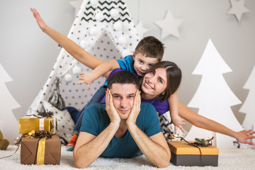Mother, father and son lying on the floor with Christmas tree and Christmas presents, play, joke, laugh, spend time together.