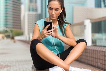 Young sporty woman resting after exercising using her smartphone and listening to music in earphones. Athlete runner in sportswear taking a break