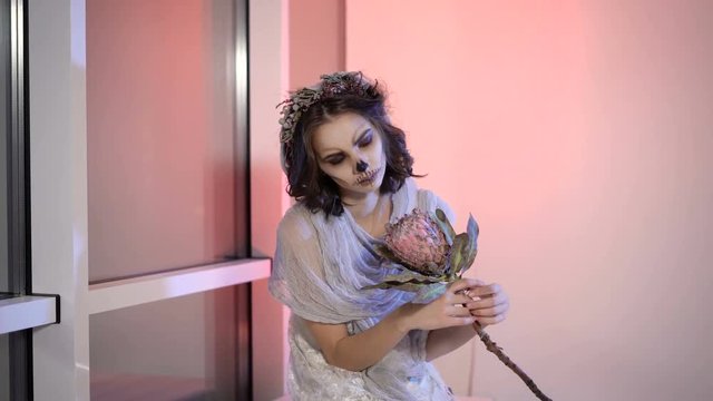 Close up of a young girl with creative scary halloween make. in appearance dead bride sitting on the bench near the window the flower in hands and looking at the camera.