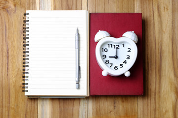 A book and notebook with a silver pencil and white alarm clock is show (9,09,21) am or pm on the wood table.