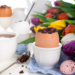 Easter egg filled with chocolate cake, tulips and colorful quail eggs on white background. Easter holidays breakfast concept.
