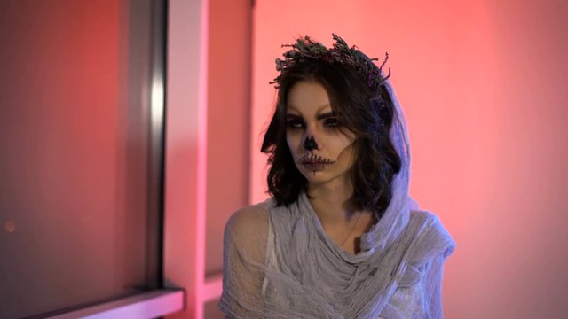 Close up of face a young girl with dead mask skull art on her . bride creative scary and creepy halloween make is sitting looking at the window. the bench the pink background.