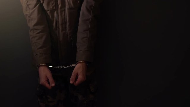 Arrested criminal cuffed man leaning on the wall in dark prison room, close up