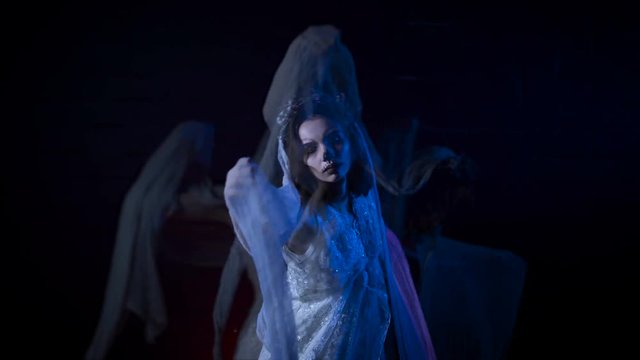 Phantom of young bride in white wedding dress and veil moving her hands. Poltergeist girl with halloween make-up is standing against black background. Mysterious woman in horror behind .