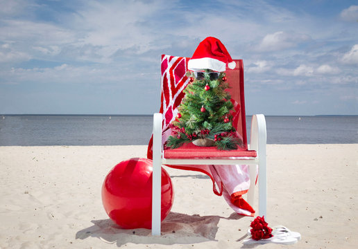 horizontal image of a green little decorated Christmas tree wearing Santa Hat and sunglasses sitting in a red lawn chair on a sandy beach with the ocean behind it on a sunny summer day.