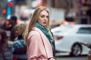 Fototapeta premium Beautiful elegant woman walking on the crowd on New York city street wearing pink fashionable jacket and scarf at winter time. Fashion model girl crossing the street.