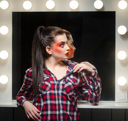 Young caucasian girl with stage make-up. A woman in a plaid shirt standing near the mirror.