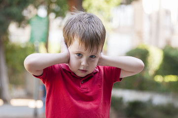 Little Caucasian boy closing ears with his hands and looking at the camera. Childhood traumatic...