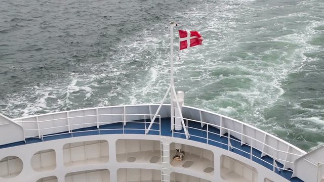 Danish flag fluttering in the wind on the stern of the ship. The ferry sails from Denmark to Sweden and back. Train on the water after the ship.